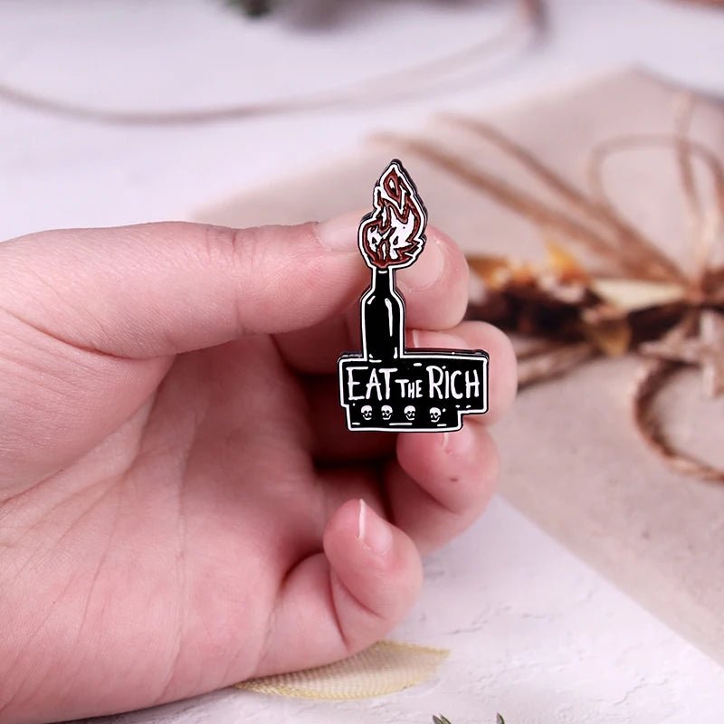 A hand displays the Eat The Rich enamel pin, shaped as a candle with a flame and cartoon skulls. Its bold black and red design contrasts against a blurred backdrop of twine-wrapped items.