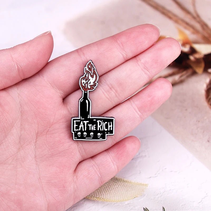 A hand holds the "Eat The Rich - Enamel Pin," featuring a flaming molotov cocktail with cartoon skulls and bold white text. The pin's black background highlights its striking design and message.