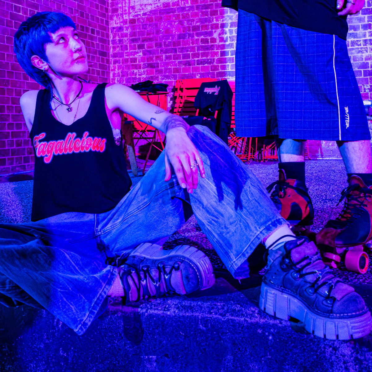 Wearing the Fagalicious locally printed satire crop tee, a person with short blue hair and chunky shoes sits on a step under purple lighting, gazing up at another individual standing in roller skates and shorts, with a brick wall and chairs in the background.
