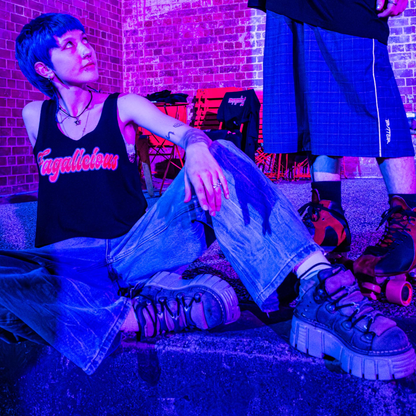 Wearing the Fagalicious locally printed satire crop tee, a person with short blue hair and chunky shoes sits on a step under purple lighting, gazing up at another individual standing in roller skates and shorts, with a brick wall and chairs in the background.