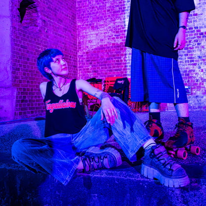 Two people wearing the Fagalicious locally printed satire crop tee and edgy outfits pose in roller skates under purple-blue lighting—one seated on concrete steps gazing upward, the other standing close by, with brick walls and chairs in the backdrop.