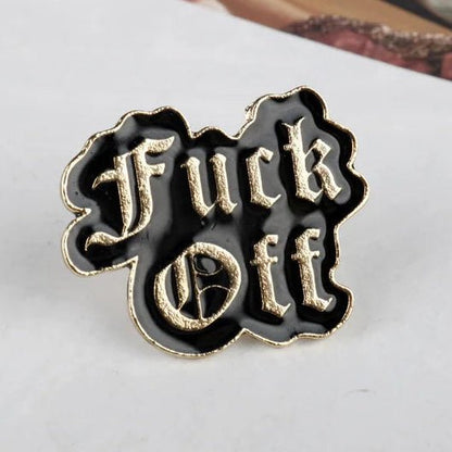 A black enamel lapel pin with gold trim and raised gothic lettering that reads 'Fuck Off'