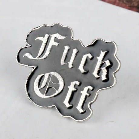 A metal enamel pin with the words 'Fuck Off' in a gothic font, featuring a black center and silver border