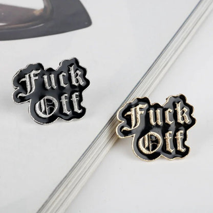 Two enamel pins with the text 'Fuck Off' in a gothic font, one with silver lettering on black and the other with gold lettering on black, displayed on a white surface.