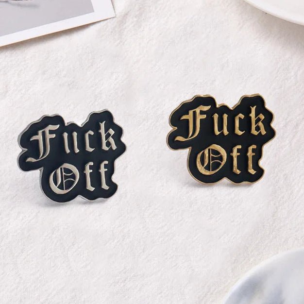 Two enamel lapel pins with the phrase 'Fuck Off' in a gothic font, one in black with gold lettering and outline, and the other in black with silver lettering and outline