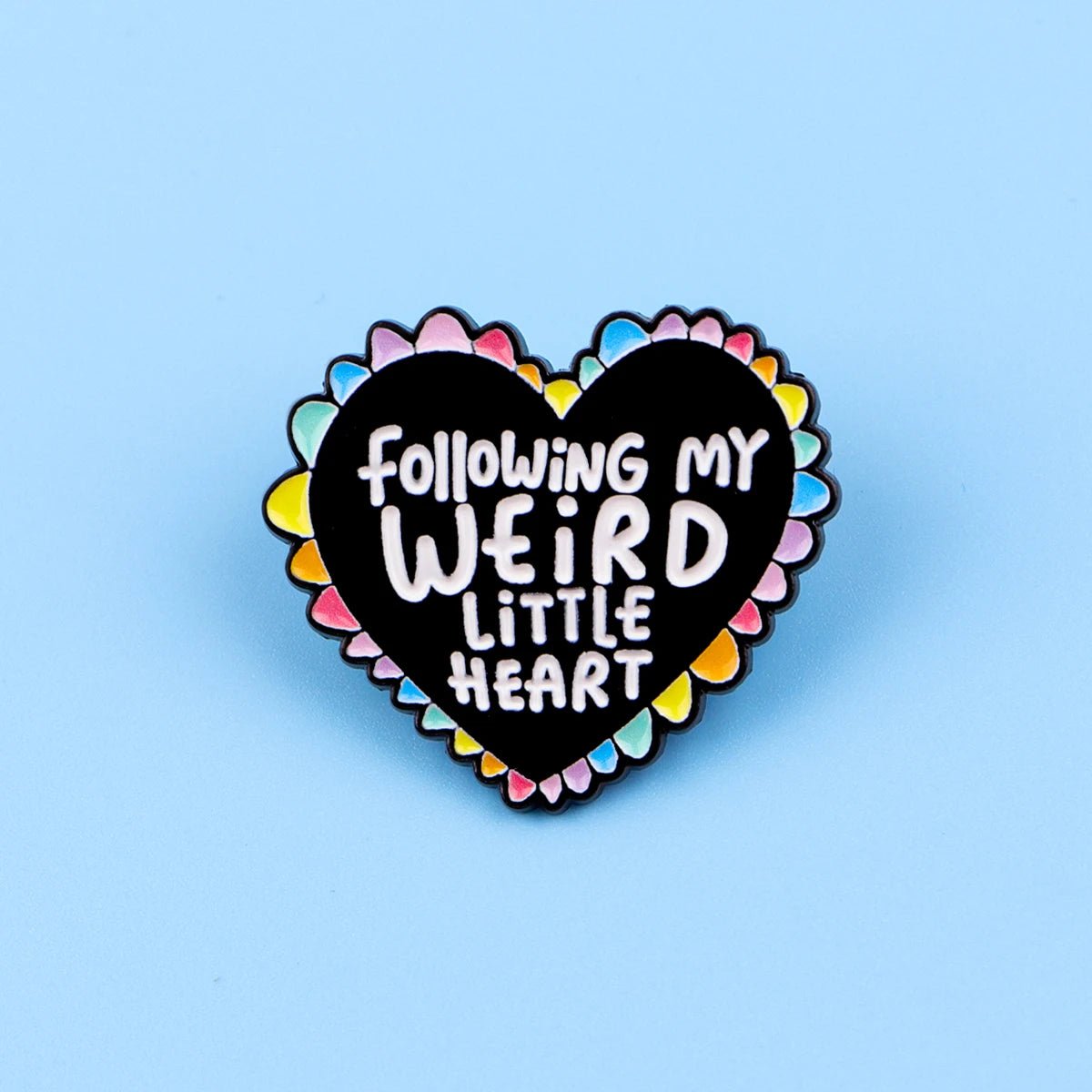 The "Following My Weird Little Heart" enamel pin features a black and rainbow heart with a colorful scalloped edge and playful white text—perfect as a pride accessory. Shown here on a blue background.