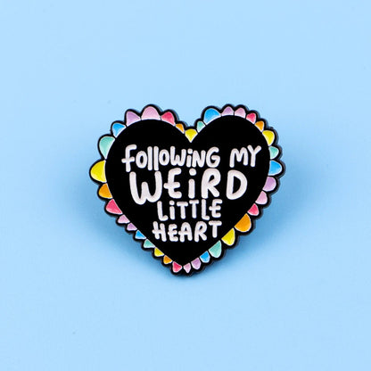 The "Following My Weird Little Heart" enamel pin features a black and rainbow heart with a colorful scalloped edge and playful white text—perfect as a pride accessory. Shown here on a blue background.