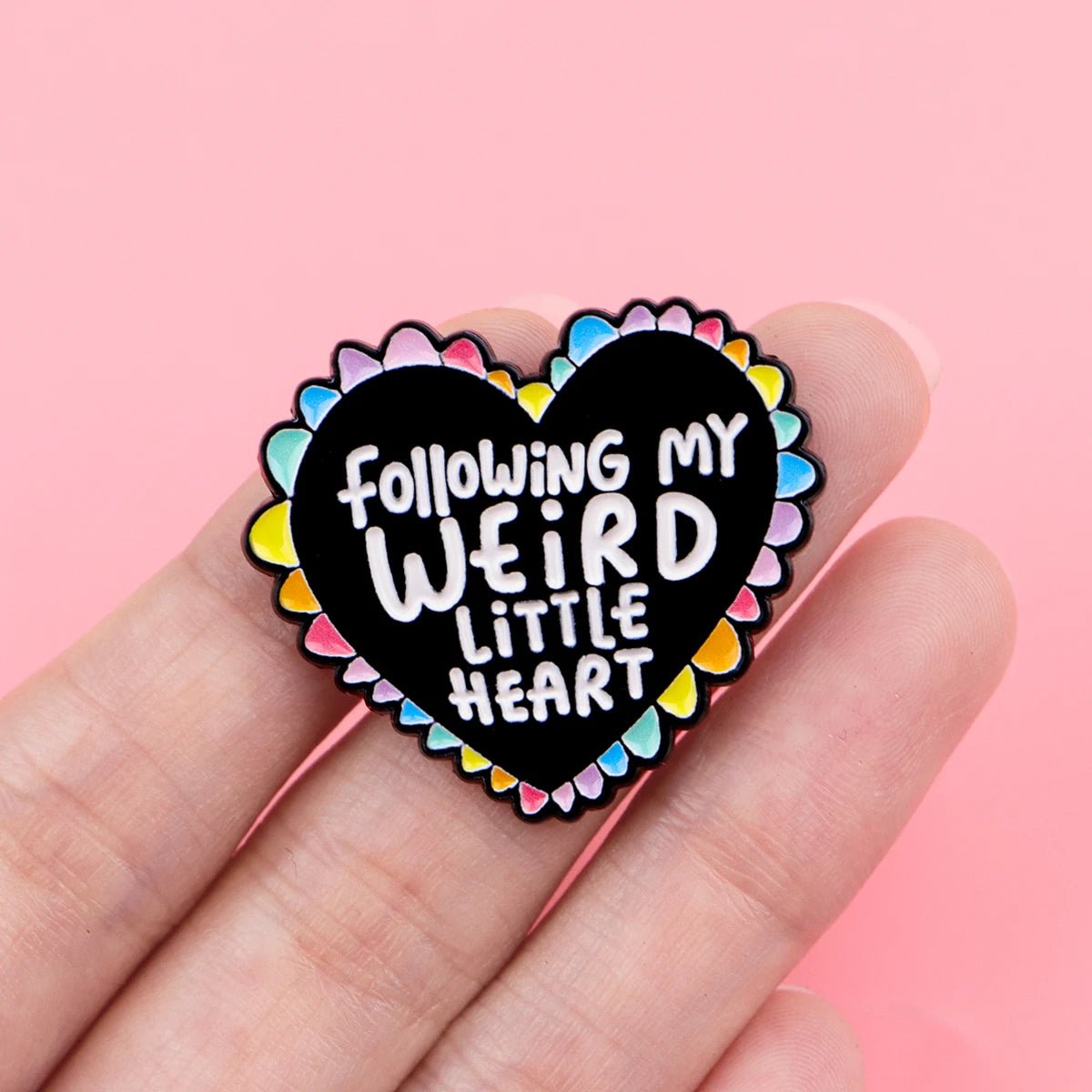 A hand displays the "Following My Weird Little Heart" enamel pin, featuring a black and rainbow heart with a scalloped colorful border and white text—an eye-catching Pride accessory.