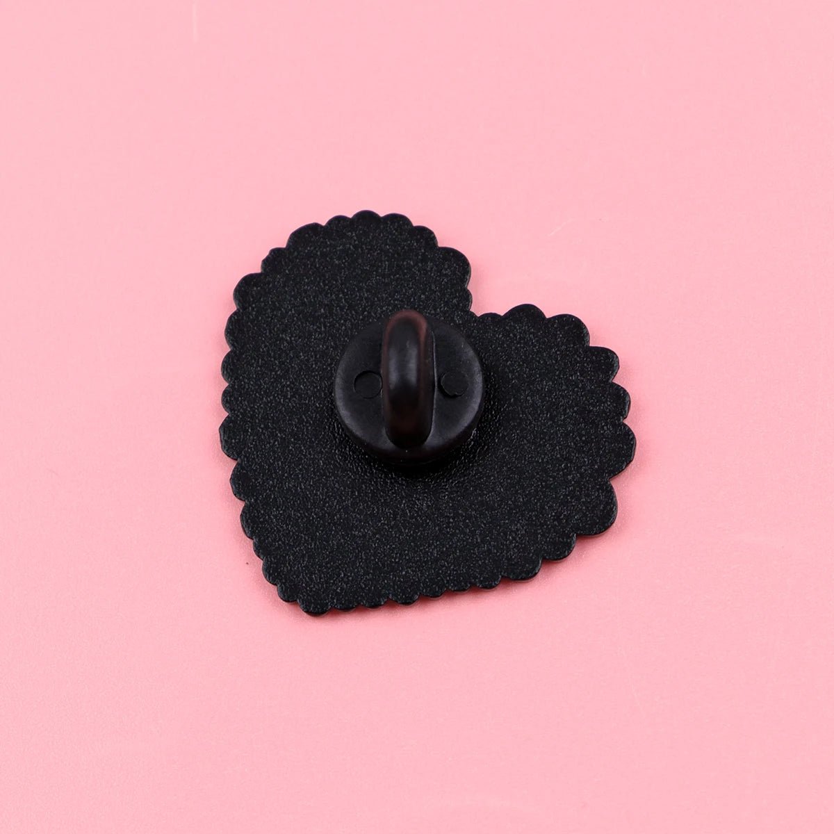 The Following My Weird Little Heart enamel pin features a black and rainbow heart with a scalloped edge and looped fastener, displayed on a solid pink background—perfect as a pride accessory.