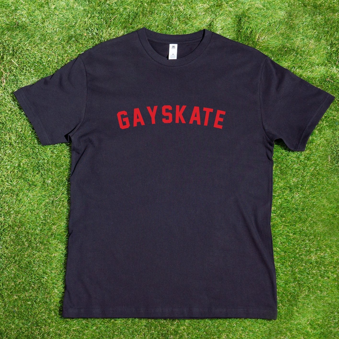 A black Gay Skate 1982 Retro T-shirt, hand-printed locally, lies flat on a grassy surface, showcasing its retro design. The shirt features the word "GAYSKATE" prominently printed in bold red capital letters across the chest. The label and tag are visible at the collar, making it ideal for custom orders.