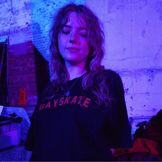A person with long wavy hair wearing a black t-shirt with 'GAYSKATE' retro printed in red across the chest, photographed in a dimly lit building with strong purple and blue ambient lighting.