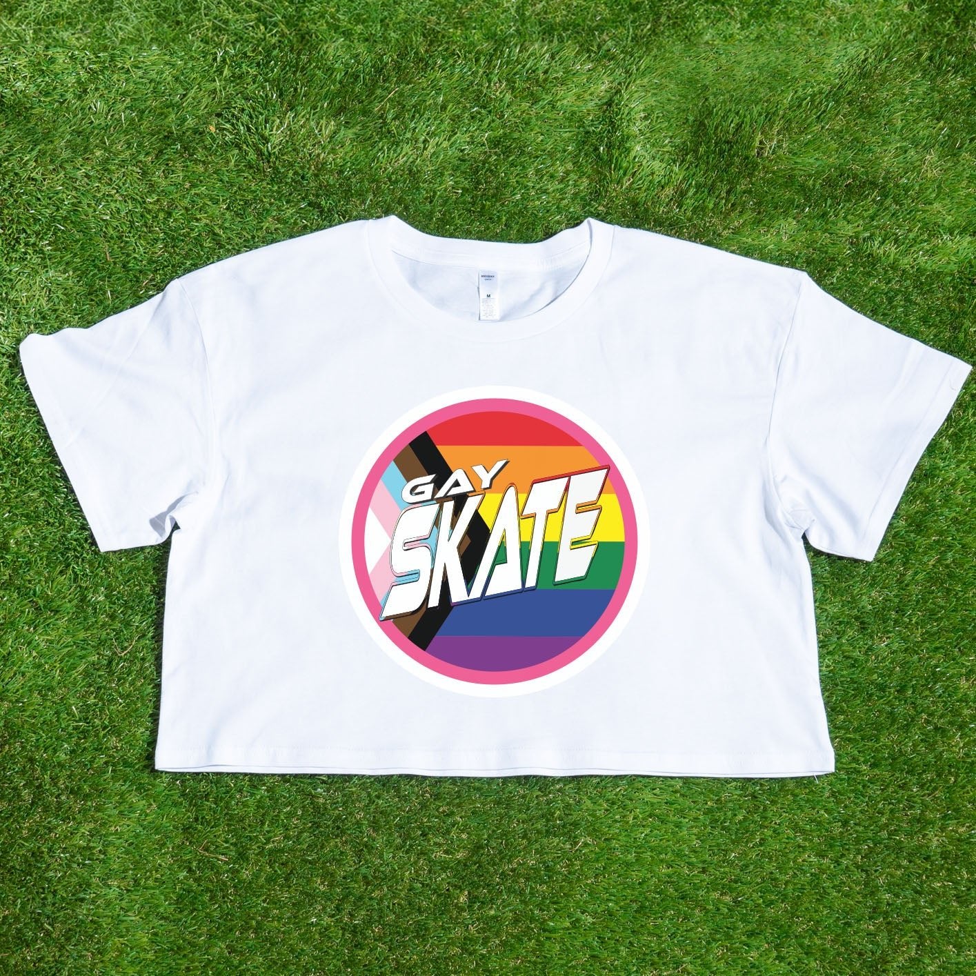 A vibrant "GAY SKATE original logo - locally printed crop tee" from Gay Skate, featuring a circular logo with the text “Gay Skates” on a rainbow background symbolizing LGBTQ+ pride, lies flat on a grassy surface. This queer skate-inspired cropped pink T-shirt exudes inclusivity and energetic spirit.