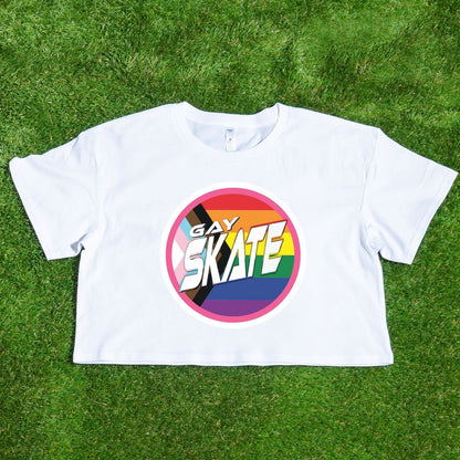 A vibrant "GAY SKATE original logo - locally printed crop tee" from Gay Skate, featuring a circular logo with the text “Gay Skates” on a rainbow background symbolizing LGBTQ+ pride, lies flat on a grassy surface. This queer skate-inspired cropped pink T-shirt exudes inclusivity and energetic spirit.