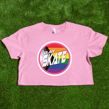 A vibrant "GAY SKATE original logo - locally printed crop tee" from Gay Skate, featuring a circular logo with the text “Gay Skates” on a rainbow background symbolizing LGBTQ+ pride, lies flat on a grassy surface. This queer skate-inspired cropped pink T-shirt exudes inclusivity and energetic spirit.