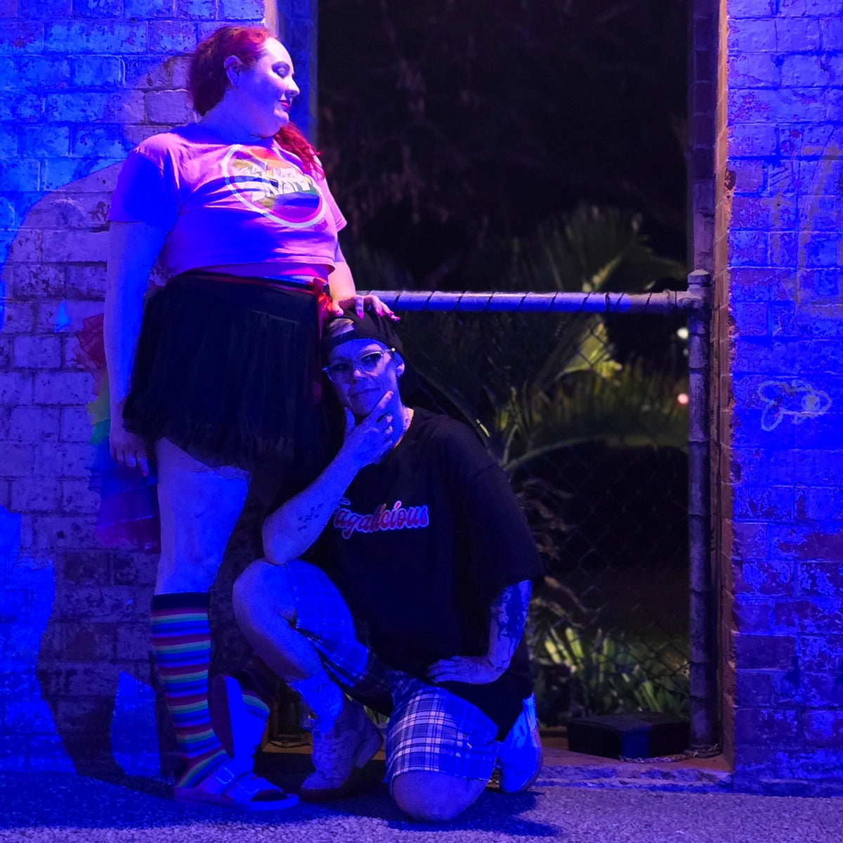 Two people pose by a brick wall illuminated with blue light. One stands wearing the GAY SKATE original logo crop tee, a black skirt, and rainbow socks. The other kneels nearby, dressed in a black shirt, plaid shorts, and a cap. Behind them are trees and a fence.