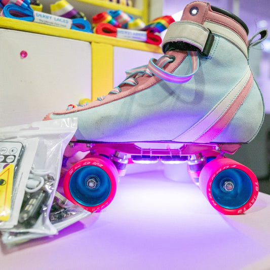 A pastel-colored roller skate with pink Hebegb wheels is illuminated by Gay Skate's vibrant  rechargeable LED skate lights. The skate lights, from the brand Gay Skate are displayed on a table beside the product which includes a small remote control, charger, and other roller skate accessories. This product is available with Australian shipping and International shipping.