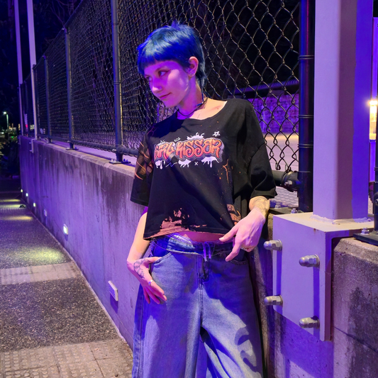 A person with short blue hair wears the “Girl Kisser” locally printed crop tee and wide-leg jeans, standing by a fence under purple lights at night, looking to the side with hands in their pockets.