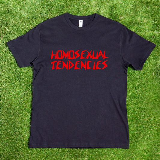 A black t-shirt from My Store, titled "HOMOSEXUAL TENDENCIES," showcases its bold, red angular font design against a lush green grass background. This locally hand-printed satire shirt embraces queer pride and celebrates diversity.