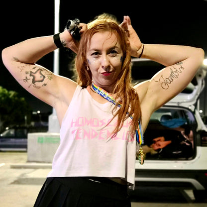 A woman with red hair confidently poses at night, revealing tattoos on her arms. She wears the “HOMOSEXUAL TENDENCIES” locally printed satire crop tee, paired with a skirt, rainbow medals, and glitter on her face. An open car trunk and lights illuminate the background.