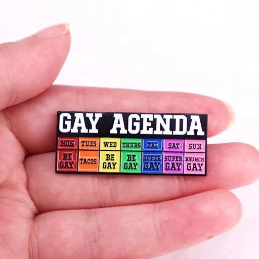 Hand holding a 'GAY AGENDA' badge with rainbow text 