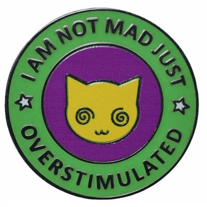 A circular enamel pin with a green outer ring reading 'I AM NOT MAD JUST OVERSTIMULATED' and a yellow cat face on a purple center, shown on a white background with a 3cm width marker below.