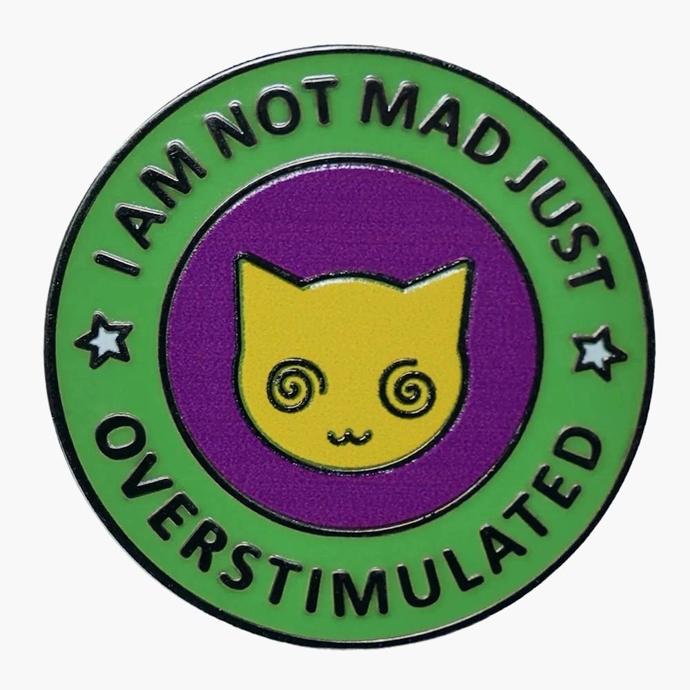 The “I Am Not Mad Just Overstimulated” enamel pin features a yellow cat face with spiral eyes on a purple background, surrounded by green edging that displays the phrase and two small stars.