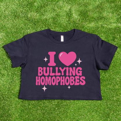 A black crop tee printed locally reads "I 🩷 Bullying Transphobes / Homophobes" in bold pink text with a heart and sparkles. Queer streetwear from a trans-owned small business, shown on green grass.