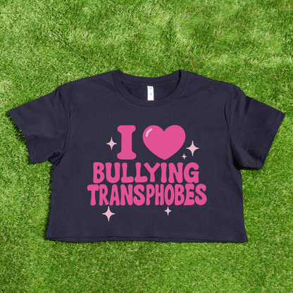 A black crop tee on grass features pink "I 🩷 BULLYING TRANSPHOBES" text with a heart and sparkles—a bold queer skate streetwear piece. Product: I 🩷 Bullying Transphobes / Homophobes - locally printed crop tee from a trans-owned business.