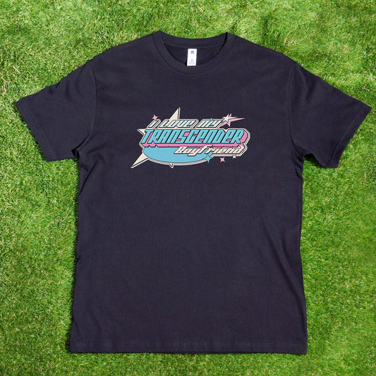 The "I LOVE MY TRANSGENDER BOYFRIEND" T-shirt from Gay Skate showcases a custom design with a vibrant retro-style graphic. The text "In Loud City, Transgender, Loud and Proud!" is prominently displayed in shades of blue, pink, and purple from the Transgender Pride Flag. The black T-shirt is laid flat on a grassy background.