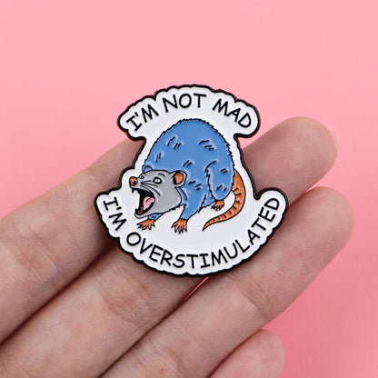 Pin with a blue rat and text 'I'm not mad, I'm overstimulated' held in a hand