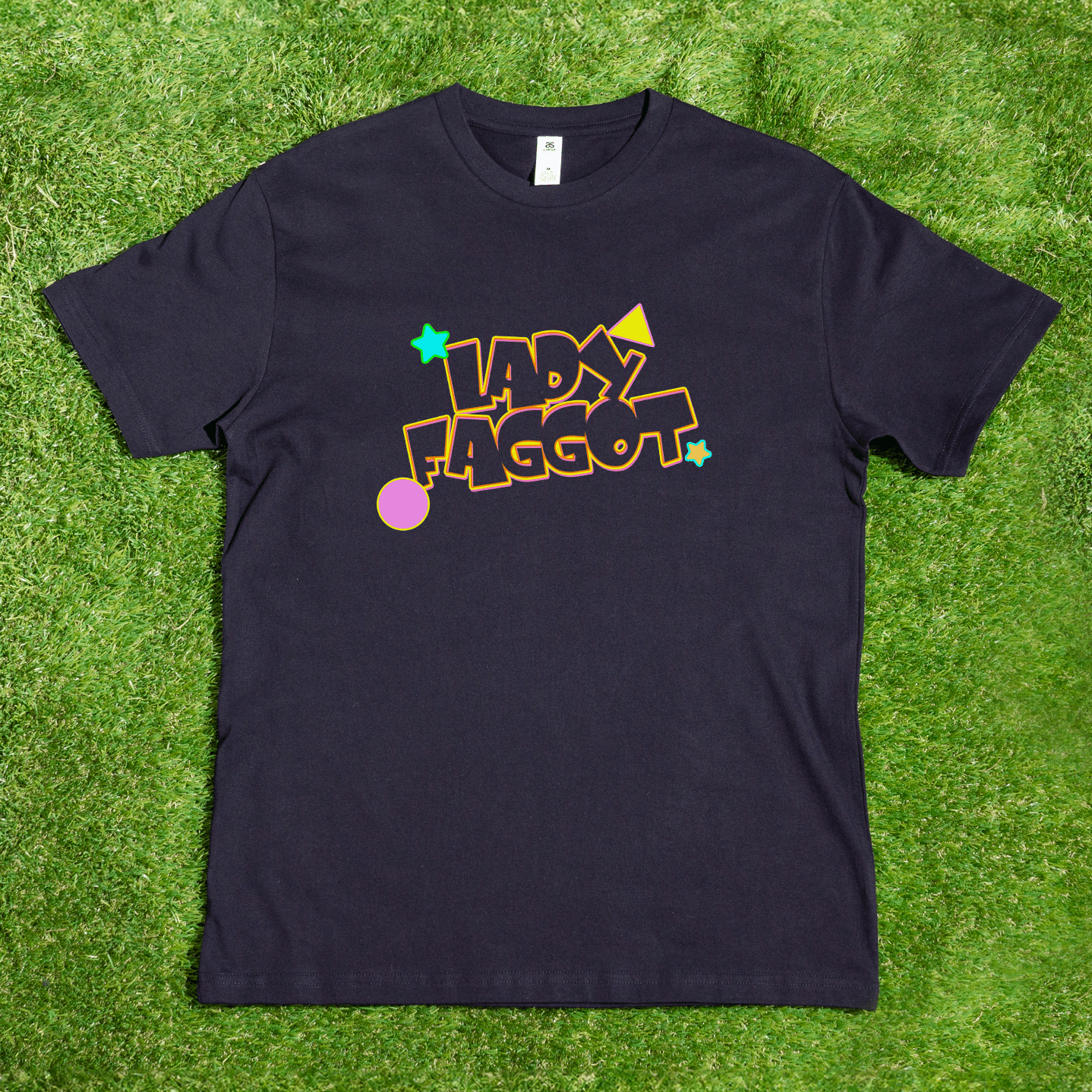 A black Lady F@gg*t - Local hand printed t-shirt lays on grass, showcasing the phrase in bold, colorful letters surrounded by stars, a circle, and geometric shapes.