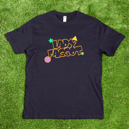 A black Lady F@gg*t - Local hand printed t-shirt lays on grass, showcasing the phrase in bold, colorful letters surrounded by stars, a circle, and geometric shapes.