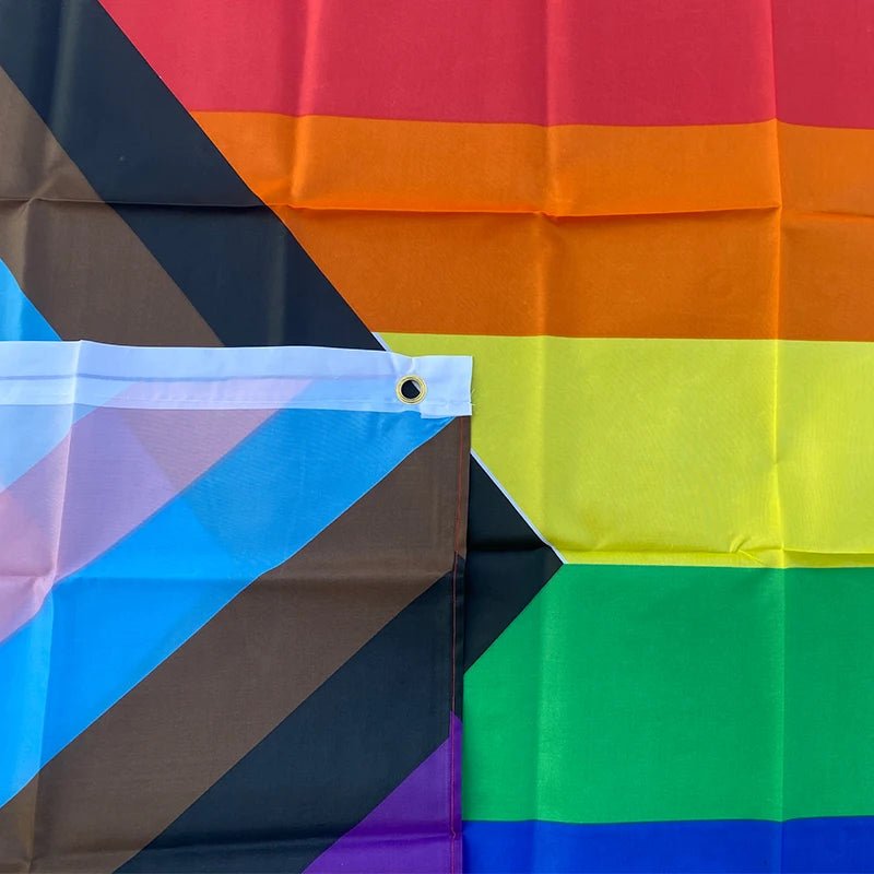 This image showcases the 150cm x 90cm Progress Pride Rainbow Flag by Gay Skate. It is made of high-quality polyester and features traditional rainbow stripes, along with a chevron on the left that includes black and brown stripes, plus light blue, white, and pink stripes to represent marginalized LGBTQ+ communities, including people of color and the trans community.