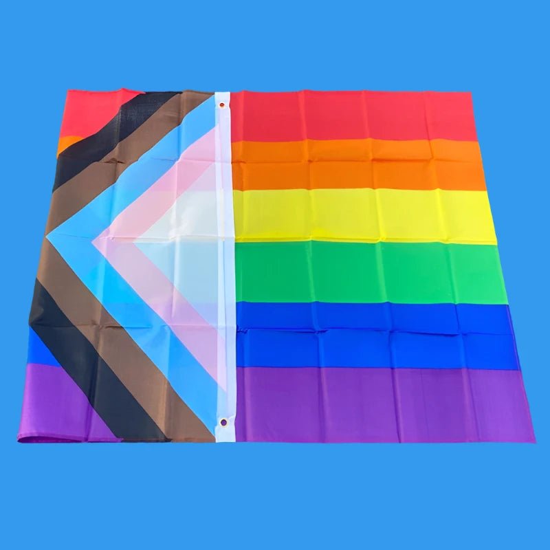 This image showcases the 150cm x 90cm Progress Pride Rainbow Flag by Gay Skate. It is made of high-quality polyester and features traditional rainbow stripes, along with a chevron on the left that includes black and brown stripes, plus light blue, white, and pink stripes to represent marginalized LGBTQ+ communities, including people of color and the trans community.