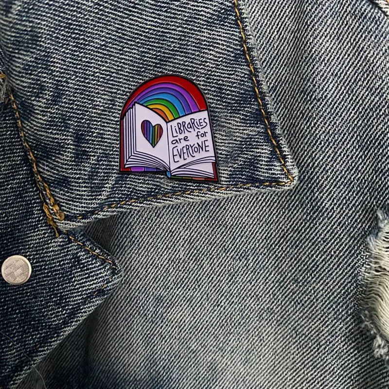 The Libraries Are For Everyone rainbow enamel pin adds a pop of color to denim, featuring an open book with a rainbow above. The text, "Libraries Are For Everyone," and heart symbol emphasize the universal love for reading.