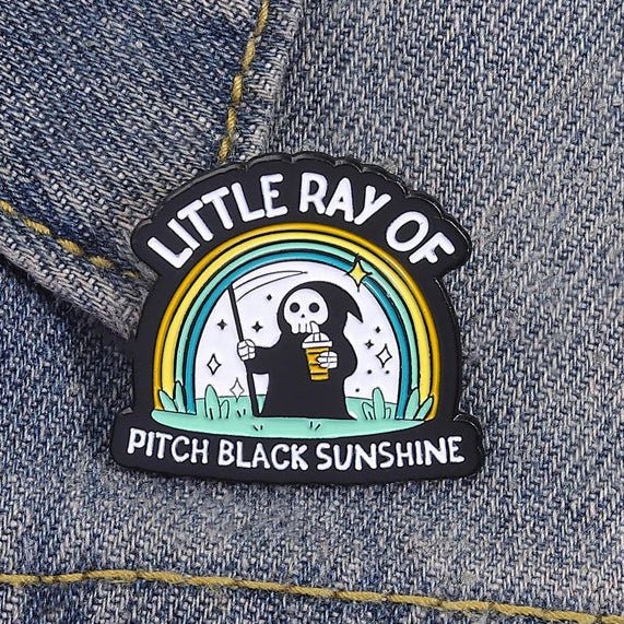 Emo Enamel pin with a design of a skeleton holding a coffee cup and rainbow, labeled 'Little Ray of Pitch Black Sunshine'.