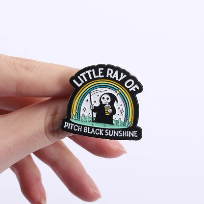 Emo Enamel pin with a design of a skeleton holding a coffee cup and rainbow, labeled 'Little Ray of Pitch Black Sunshine'.