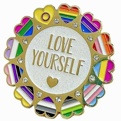 The "Love Yourself - ultra pride enamel pin" features gold "LOVE YOURSELF" lettering at the center, surrounded by heart-shaped multi-colored pride flags and sparkling rhinestones on a round gold border.