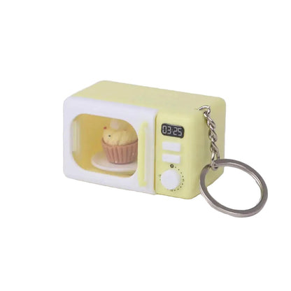 A miniature yellow and white microwave keychain with a small cupcake figurine inside and a metal key ring attached, shown on a clean light gray background.