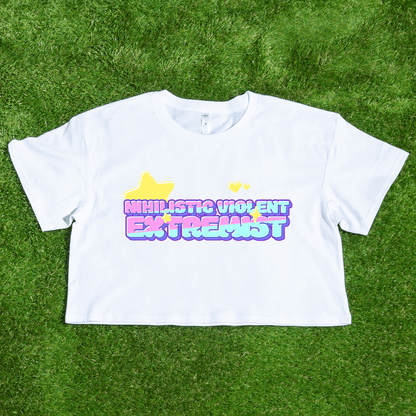 The "Nihilistic Violent Extremist" locally printed satire crop tee features pastel bubble text, yellow hearts and stars on a white shirt. Hand-printed by a trans and queer owned brand and shown displayed on green grass.