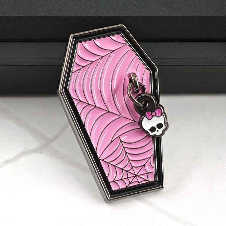 A monster high coffin-shaped pink enamel pin featuring a cobweb pattern with a small Hello Kitty styleskull charm