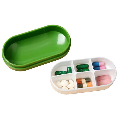 A green oval pill organizer tray shown open with stacked green lids, alongside a white divided compartment tray filled with assorted capsules and tablets on a beige surface.