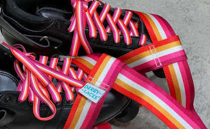 A pair of black roller skates with durable Waxed Skate Laces in lesbian pride flag colors and a matching "Derby Laces" strap rest on a gray surface.