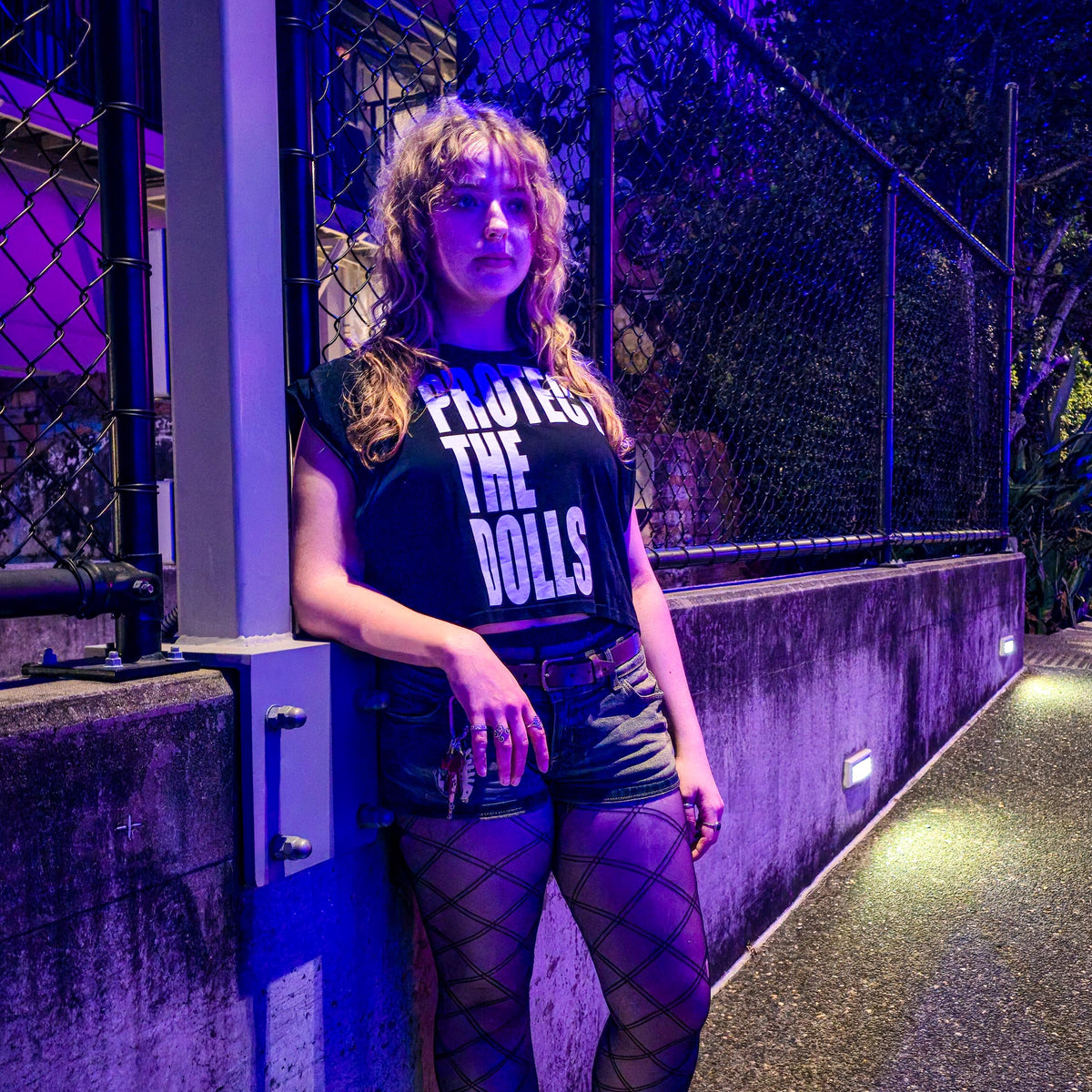 A person with wavy hair stands by a fence at night under purple lighting, wearing the Protect The Dolls crop tee, shorts, fishnet tights, and holding a drink.