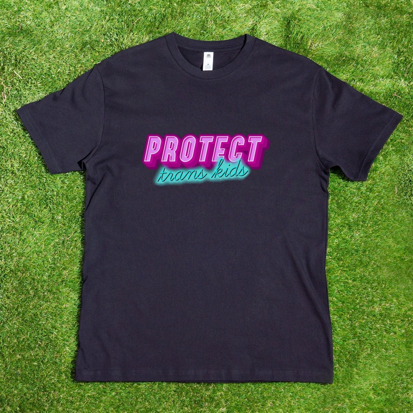 The "Protect Trans Kids" - Local hand printed t-shirt by Gay Skate is a black AS Colour shirt showcasing the phrase in eye-catching pink and blue lettering, echoing the colors of the Transgender Flag, and elegantly set against a vibrant green grass backdrop.