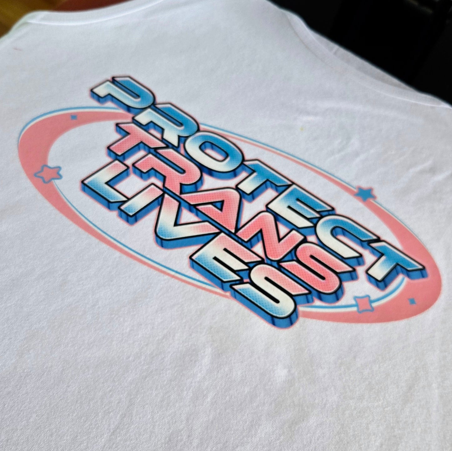 The "Protect Trans Lives - Local hand printed t-shirt" features a white base with the message "Protect Trans Lives" in vibrant, colorful letters. A blue and pink elliptical design with stars reflects diversity and transgender pride colors.