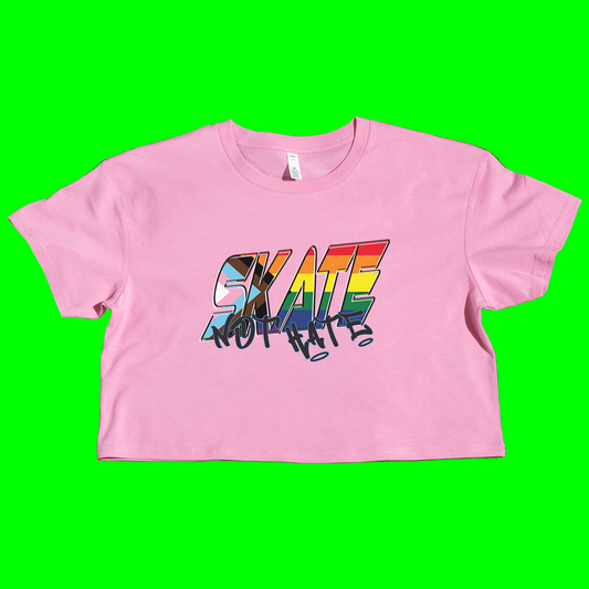 A white cropped t-shirt with the text 'SKATE NOT HATE' printed in rainbow colors on the front, placed on a grassy surface.
