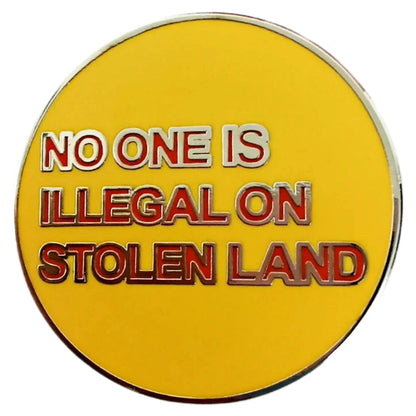 A round yellow enamel pin, titled “No One Is Illegal On Stolen Land,” features the phrase in bold red and white letters.