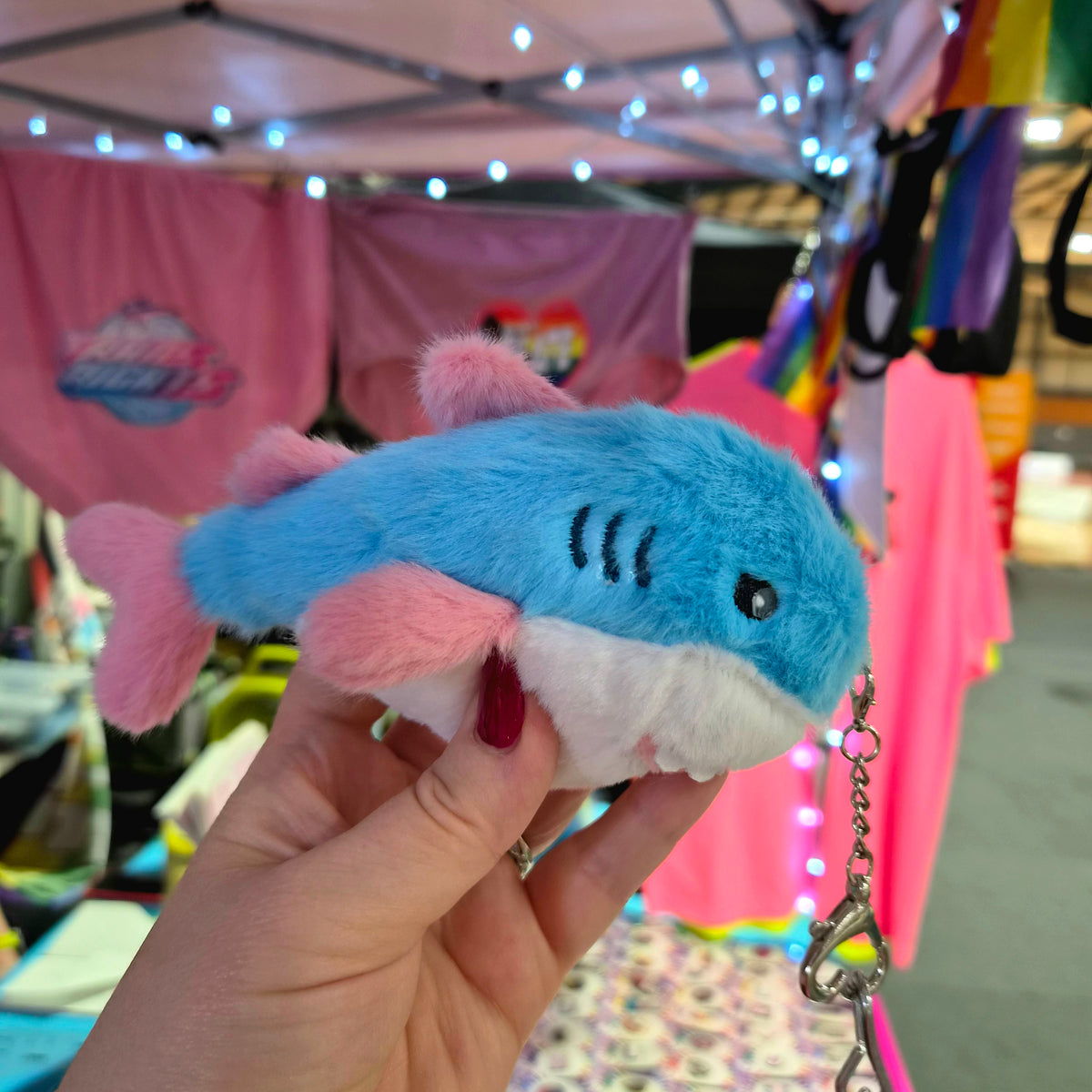 A hand holds a Trans Pride Plush Shark Keychain - 15cm BLAHAJ-Inspired Heart Carabiner Clip-On Bag, featuring a small plush shark patterned in blue, white, and pink with a metal heart-shaped carabiner.