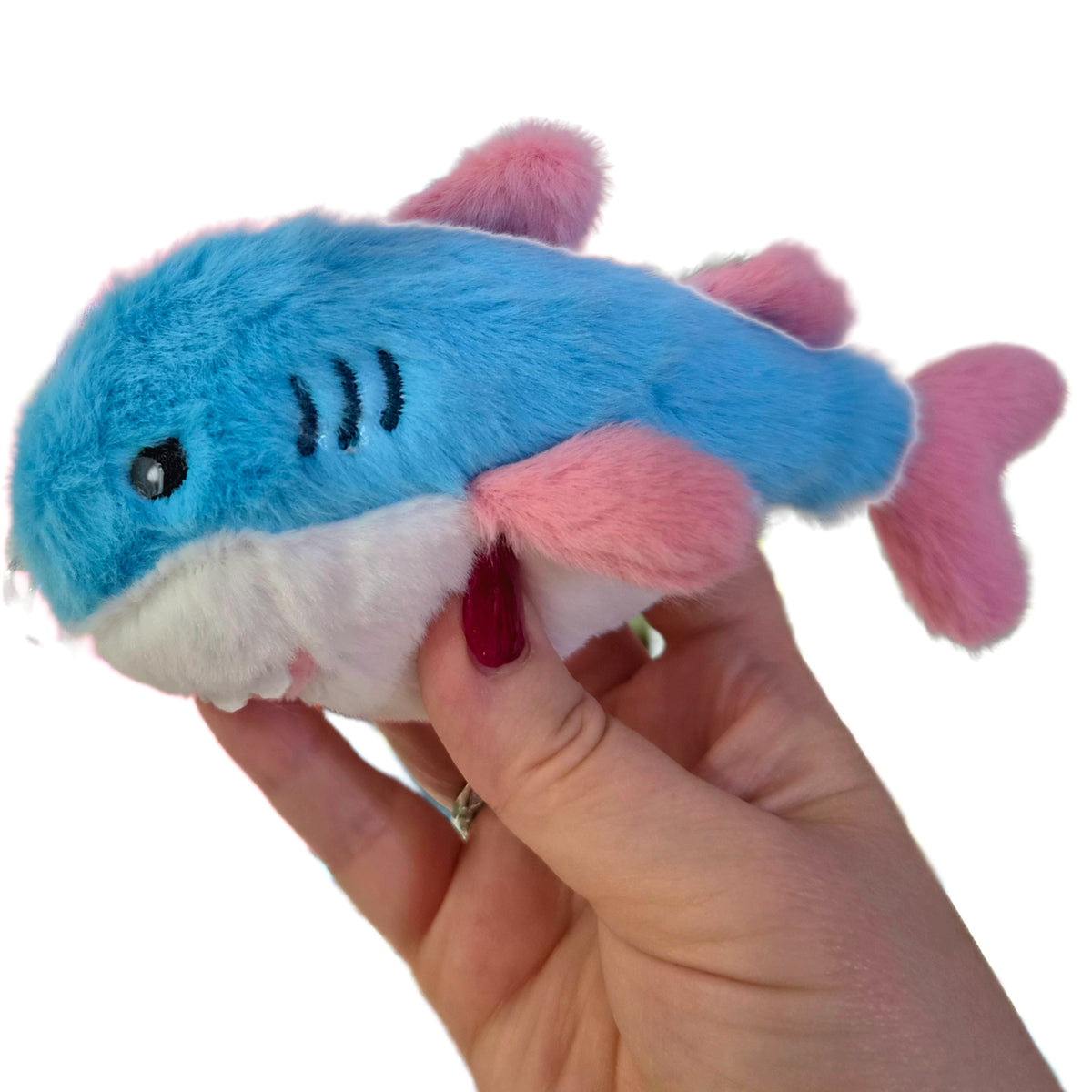 A hand holds a Trans Pride Plush Shark Keychain - 15cm BLAHAJ-Inspired Heart Carabiner Clip-On Bag, which is a small plush shark with a blue body, white belly, and pink fins.